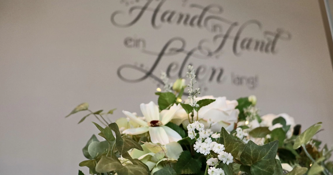 Im Vordergrund weißer Blumenschmuck, im Hintergrund der Schriftzug "Hand in Hand ein Leben lang" an der Wand.