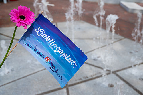 Eine blaue Papierflagge mit der Aufschrift Lieblingsplatz und eine Blume werden vor das Wasserspiel auf dem La Couronne-Platz gehalten