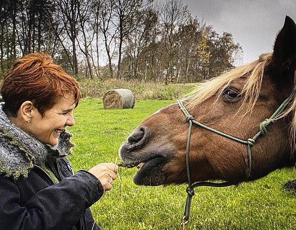 Reitpädagogin Mona Pelz füttert ein Pferd mit Gras. Im Hintergrund eine grüne Wiese.