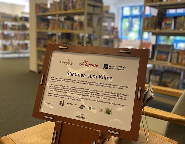Ein Informationsschild mit dem Ausstellungstitel "Stimmen zum Klima" steht auf einem Hocker. Im Hintergrund Bücherregale.