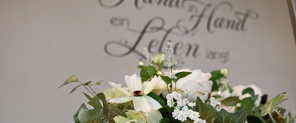Im Vordergrund weißer Blumenschmuck, im Hintergrund der Schriftzug "Hand in Hand ein Leben lang" an der Wand.