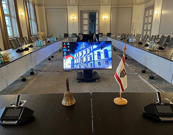 Ständesaal mit den im Rund gestellten Tischen, in der Mitte stehen Monitore auf denen ein Bild vom Historischen Rathaus zu sehen ist. 