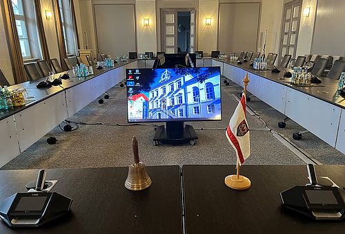 Ständesaal mit den im Rund gestellten Tischen, in der Mitte stehen Monitore auf denen ein Bild vom Historischen Rathaus zu sehen ist. 
