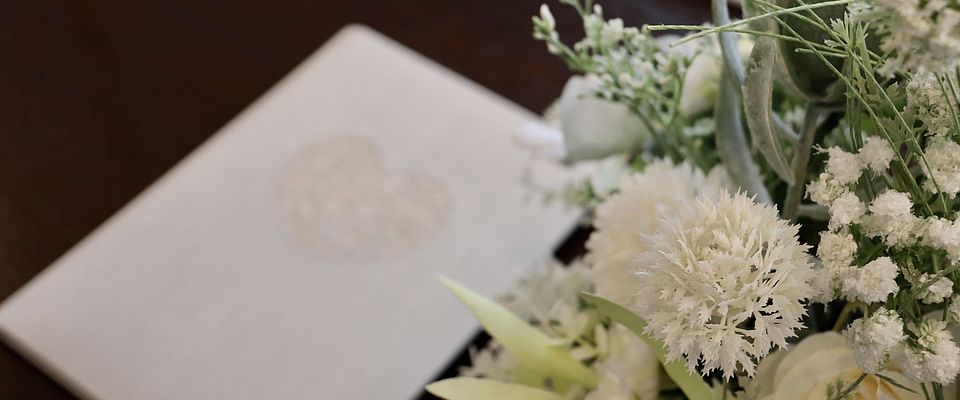 Rechts im Vordergrund weißer Blumenschmuck. Verschwommen im Hintergrund liegt eine weiße Karte mit Herzmotiv auf dem Tisch.