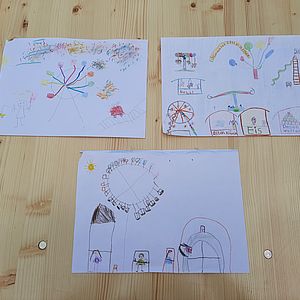 Drei von Kindern gemalte Bilder zum Thema Jahrmarkt auf einem Tisch. Die Bilder zeigen verschiedene Fahrgeschäfte und Buden.